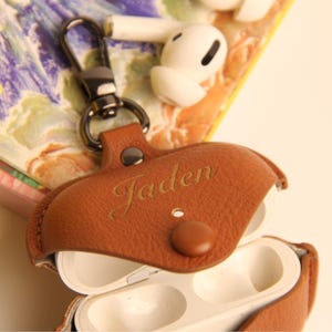 May include: A brown leather AirPod case with the name "Jaden" engraved on the front. The case is open, revealing white AirPods inside. A black carabiner clip is attached to the case. The case is resting on a floral patterned surface.