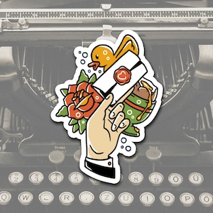 May include: A sticker showing a hand holding a letter sealed with a heart, a rose, a duck, and a green and brown cup. The sticker is set against a vintage typewriter.