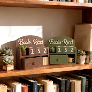 May include: Two wooden book counters with the words "Books Read" in white script. One is brown with a drawer and a small plant, the other is green with a pumpkin and a small drawer. Both show the number 152.