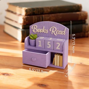May include: A lavender-colored wooden bookshelf with the words "Books Read" on top. It features a small drawer, a miniature succulent, and numbered blocks displaying "152". The bookshelf is 5.5 inches wide, 5.3 inches tall, and 1.2 inches deep.