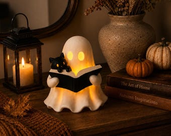 May include: A cream-colored ghost figurine with glowing yellow eyes, holding an open black book with a small black cat perched on the edge. The ghost is illuminated from within, casting a warm light on the surrounding surface and objects.