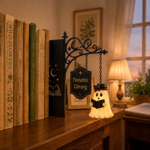 Könnte beinhalten: Ein Buchstütze aus schwarzem Metall mit einer dekorativen Schnörkelhalterung und einem hängenden Schild mit der Aufschrift Pamela's Library. Eine kleine, leuchtend weiße Geisterfigur mit einer schwarzen Katze auf dem Kopf hält ein offenes schwarzes Buch.
