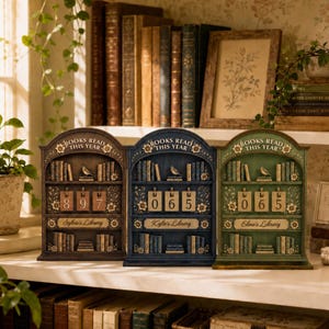 May include: Three arched wooden book trackers in brown, blue, and green, each featuring the text 'BOOKS READ THIS YEAR' at the top, a shelf with miniature book carvings, a bird figure, and a central flip-style counter for tracking reading progress.