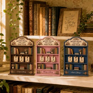 May include: Three wooden bookshelf-shaped trackers labeled BOOKS READ THIS YEAR, available in brown, pink, and blue. Each features two shelves with miniature book decorations, a central row of three numbered date cards, and a personalized name plate.