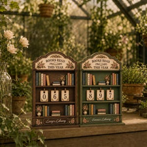 May include: Two wooden bookshelf-shaped trackers labeled BOOKS READ THIS YEAR with floral accents. One is brown with Lucy's Library and the number 065, the other is green with Emma's Library and the number 897. Both feature miniature book spines.