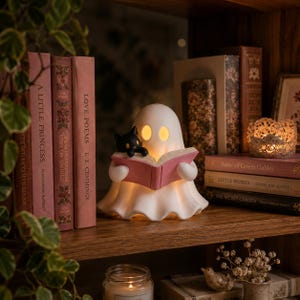 May include: A white ghost-shaped lamp with glowing yellow eyes, holding an open pink book with a small black cat perched on the edge. The lamp is positioned on a wooden shelf between several hardcover books, including A Little Princess and Love Poems.