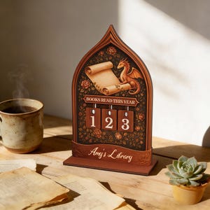 Może przedstawiać: Drewniany licznik książek z motywem smoka i zwoju. Licznik wyświetla napis "BOOKS READ THIS YEAR" i liczbę "123". Na podstawie napis "Amy's Library". Licznik jest brązowy z kwiatowymi akcentami i ilustracją smoka.