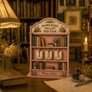 May include: A pink wooden bookshelf display with three shelves, featuring the text BOOKS READ THIS YEAR at the top and Mary's Library at the bottom. The shelves hold miniature books, a small floral vase, and three hanging tags numbered 1, 2, and 3.