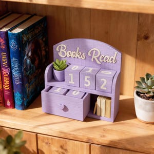 May include: A lavender wooden desk organiser with the words "Books Read" in white script. The organiser has a small drawer, a shelf with small books, and numbered blocks displaying "152". A small potted succulent sits on the top shelf.