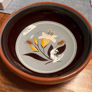 Puede incluir: Un cuenco redondo de cerámica con exterior e interior marrón. El interior presenta un centro azul claro con un diseño floral en blanco, amarillo y marrón. El borde es de color naranja cálido.