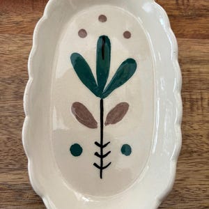 May include: Cream-colored ceramic oval dish with a scalloped edge. The dish features a hand-painted floral design in green, brown, and black. The design includes a central stem with leaves and dots. The dish is on a wooden surface.