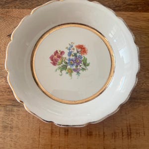 Puede incluir: Un plato blanco con borde festoneado, borde dorado y diseño floral en el centro. Las flores son una mezcla de rojo, morado, azul y naranja. El plato es probablemente de porcelana.