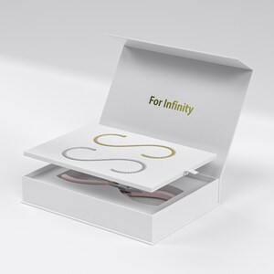 Może przedstawiać: Białe pudełko prezentowe z napisem "For Infinity" w złotych literach. Wewnątrz biała taca z dwoma bransoletkami w kształcie litery S, jedną złotą i jedną srebrną. Poniżej w pudełku spoczywają różowe szczypce.