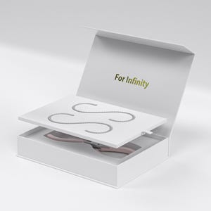 Puede incluir: Una caja de regalo blanca con el texto "For Infinity" en letras doradas. En el interior, una bandeja blanca contiene tres objetos plateados curvos en forma de cadena. Debajo, se ve una herramienta rosa. La caja está abierta.