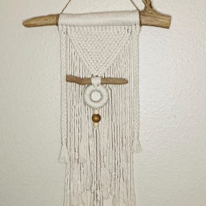 May include: Cream-colored macrame wall hanging with a natural wood branch at the top. The design features a geometric pattern, a wooden ring, and a wooden bead. The bottom has multiple tassels, creating a bohemian aesthetic.