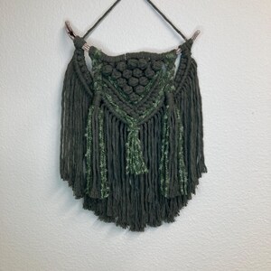 Puede incluir: Colgante de pared de macramé hecho a mano en tonos verdes. El diseño presenta un patrón geométrico central con nudos texturizados y flecos. La pieza se suspende de un pasador de madera con un cordón para colgar, creando una estética bohemia.