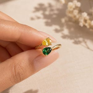 May include: A gold bypass ring featuring two pear-cut gemstones, one yellow and one green, set in a polished metal band. The open design allows the stones to sit opposite each other, creating a distinct contrast between the vibrant yellow and deep green colours.