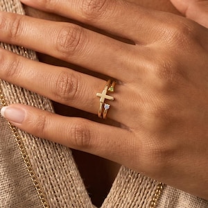 May include: A gold ring set featuring a cross design and a smaller ring with a gemstone. The rings are worn on a finger, with a neutral-toned background. The jewelry is simple and elegant.