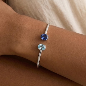 May include: A silver cuff bracelet with two oval gemstones: a dark blue sapphire and a light blue aquamarine. The open design allows the bracelet to be easily worn. The gemstones are set in a minimalist style. The bracelet is a piece of jewellery.