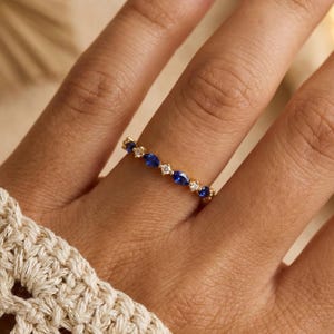 May include: A gold band ring featuring alternating oval blue gemstones and small round clear gemstones. The ring is set on a finger, with a cream-colored crocheted sleeve in the foreground. The ring's design is delicate and elegant.