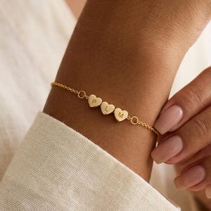 May include: A gold-colored bracelet featuring four heart-shaped charms, each with a letter: P, L, M. The bracelet is delicate and worn on a wrist, with a white shirt in the background. The bracelet is a personalized jewelry item.
