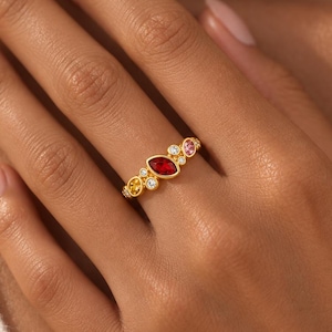Peut inclure: Une bague en or ornée d'une pierre rouge centrale taille marquise, entourée de petites pierres rondes et ovales dans des tons jaune, rose et blanc transparent. L'anneau est en or jaune poli, mettant en valeur un groupe de pierres multicolores.
