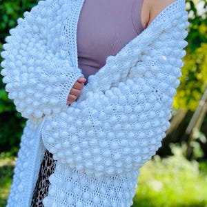 May include: A white, longline cardigan with a textured bobble stitch pattern. This handmade, oversized sweater is a comfortable and fashionable layering piece. The cardigan is a soft, off-white colour.