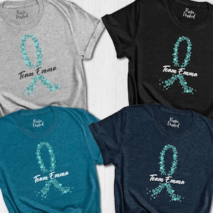 May include: Four t-shirts in gray, black, teal, and navy blue. Each shirt features a teal ribbon design made of small hearts, with the words "Team Emma" in a script font below the ribbon. The shirts are short-sleeved.