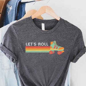 May include: Dark gray t-shirt with the text "LET'S ROLL" and a retro roller skate graphic. The graphic features a colorful stripe design in orange, yellow, and green. The roller skate is teal and orange.