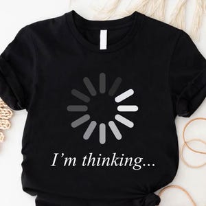 May include: Black t-shirt featuring a white and gray loading circle graphic. Below the graphic, the text reads "I'm thinking...". The shirt is made of a soft material and has short sleeves.