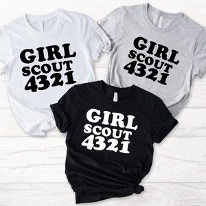 Puede incluir: Tres camisetas de manga corta en blanco, gris jaspeado y negro. Cada camiseta presenta el texto GIRL SCOUT 4321 impreso en una fuente negrita y redondeada. Las camisetas blanca y gris tienen texto negro, mientras que la negra tiene texto blanco.