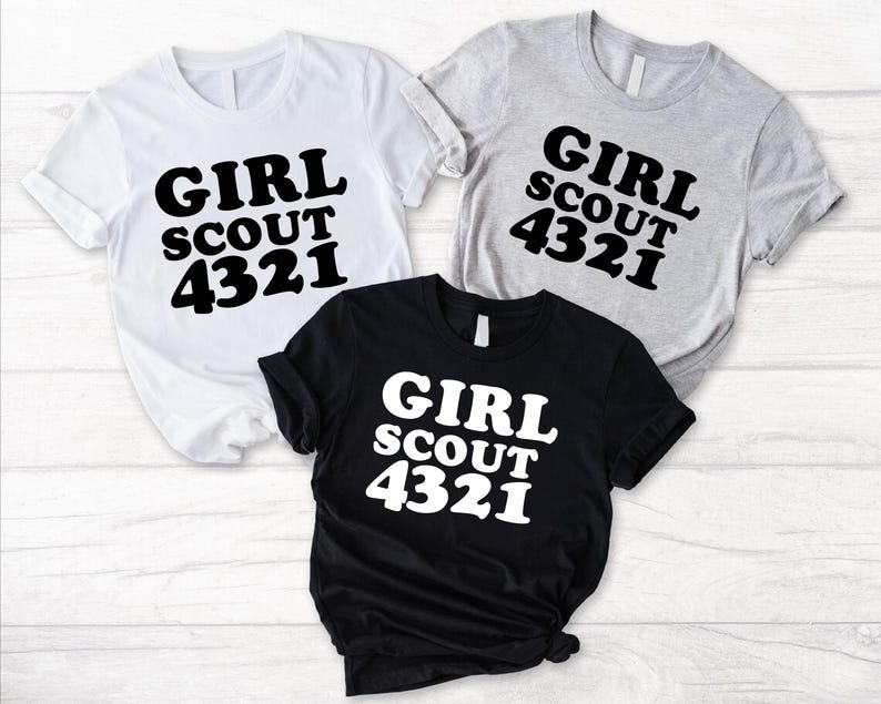 Puede incluir: Tres camisetas de manga corta en blanco, gris jaspeado y negro. Cada camiseta presenta el texto GIRL SCOUT 4321 impreso en una fuente negrita y redondeada. Las camisetas blanca y gris tienen texto negro, mientras que la negra tiene texto blanco.