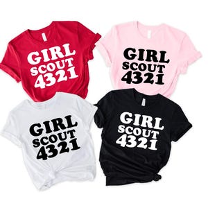 Puede incluir: Cuatro camisetas de manga corta en rojo, rosa claro, blanco y negro. Cada camiseta presenta el texto "GIRL SCOUT 4321" impreso en una fuente negrita y redondeada. Las camisetas roja y blanca tienen texto blanco, y las rosa y negra tienen texto negro.