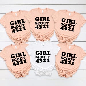 Puede incluir: Cinco camisetas de manga corta en color melocot&oacute;n claro y una en blanco, cada una con el texto negro GIRL SCOUT 4321 impreso en el frente. Las camisetas est&aacute;n dispuestas en dos filas, con cada una atada en un nudo en el dobladillo inferior.