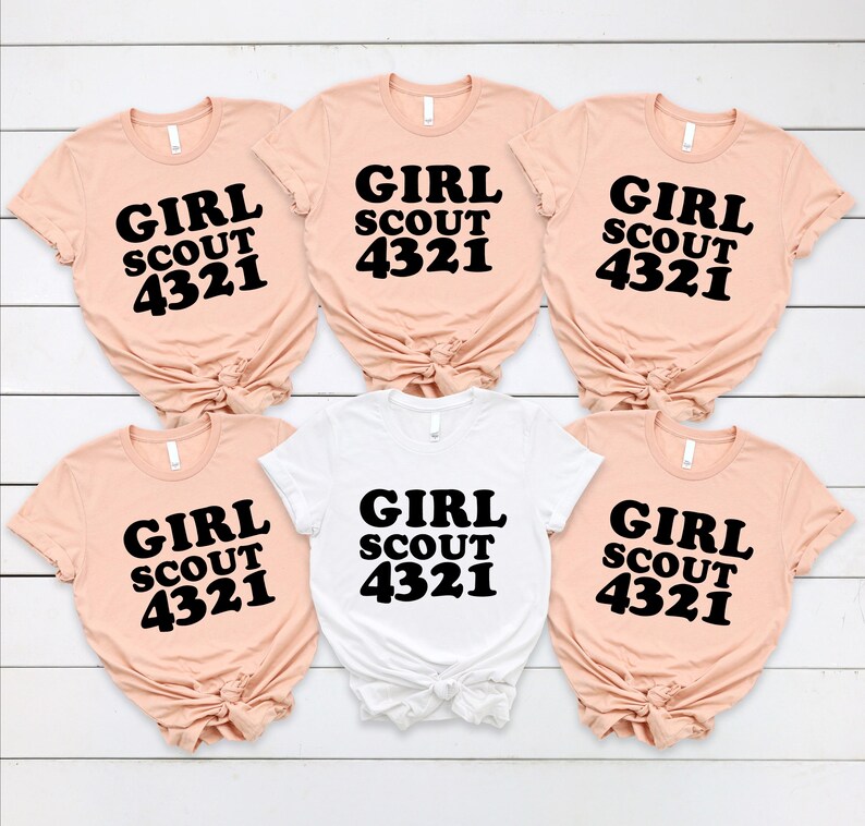 Puede incluir: Cinco camisetas de manga corta en color melocot&oacute;n claro y una en blanco, cada una con el texto negro GIRL SCOUT 4321 impreso en el frente. Las camisetas est&aacute;n dispuestas en dos filas, con cada una atada en un nudo en el dobladillo inferior.