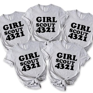 Puede incluir: Cinco camisetas de manga corta de color gris claro dispuestas en grupo. Cada camiseta presenta el texto negro "GIRL SCOUT 4321" impreso en una fuente negrita y redondeada en el pecho. Las camisetas tienen un cuello redondo est&aacute;ndar.