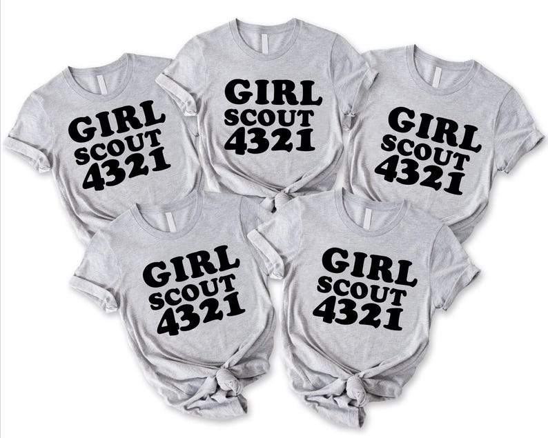 Puede incluir: Cinco camisetas de manga corta de color gris claro dispuestas en grupo. Cada camiseta presenta el texto negro "GIRL SCOUT 4321" impreso en una fuente negrita y redondeada en el pecho. Las camisetas tienen un cuello redondo est&aacute;ndar.