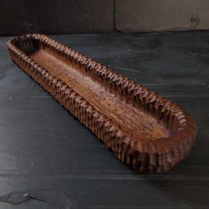 May include: A long, hand-carved wooden tray with a textured, ridged exterior and a smooth interior. The tray is made of dark brown wood and has an oval shape. The tray is suitable for serving food or displaying decorative items.