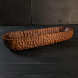 May include: A hand-carved wooden bowl with a textured surface. The bowl is oval-shaped and has a dark brown colour. The interior of the bowl is smooth, while the exterior features a carved pattern. This decorative bowl is suitable for serving food or as a decorative item.