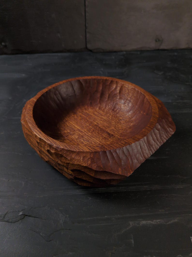 May include: A hand-carved wooden bowl in a deep reddish-brown colour. The bowl has a textured, organic design with carved ridges around the outside. The interior is smooth, contrasting with the rough exterior. A unique, rustic decorative piece.