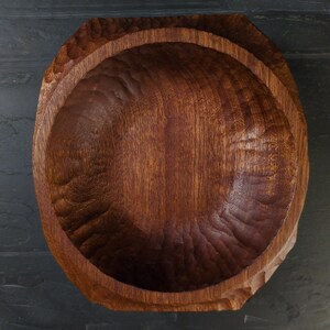 May include: A hand-carved wooden bowl in a deep, reddish-brown hue. The interior features a textured surface, while the exterior has a faceted design. The visible wood grain enhances its natural appeal. This bowl is suitable for serving or as a decorative item.