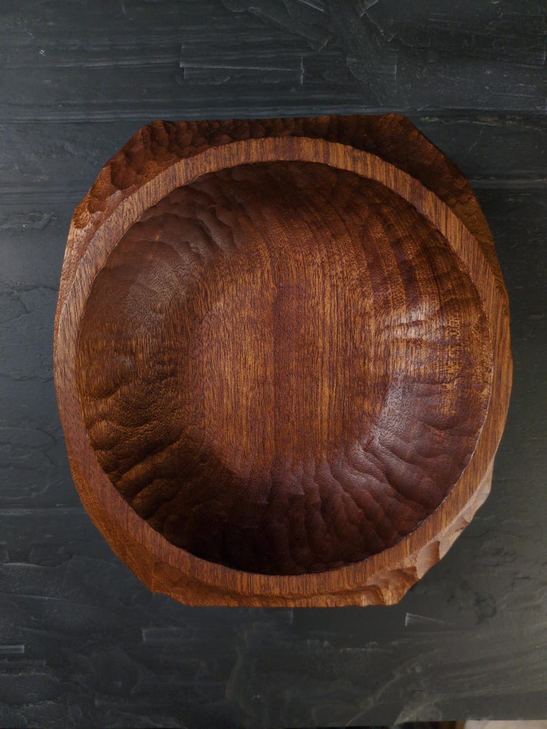 May include: A hand-carved wooden bowl in a deep, reddish-brown hue. The interior features a textured surface, while the exterior has a faceted design. The visible wood grain enhances its natural appeal. This bowl is suitable for serving or as a decorative item.