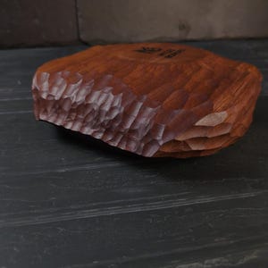 May include: A hand-carved wooden bowl with a textured, chiseled edge. The bowl is a warm reddish-brown colour and appears to be made from a single piece of wood. The interior of the bowl is smooth, and the exterior has a rough, organic texture.