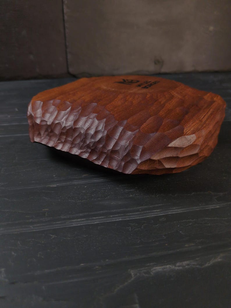 May include: A hand-carved wooden bowl with a textured, chiseled edge. The bowl is a warm reddish-brown colour and appears to be made from a single piece of wood. The interior of the bowl is smooth, and the exterior has a rough, organic texture.