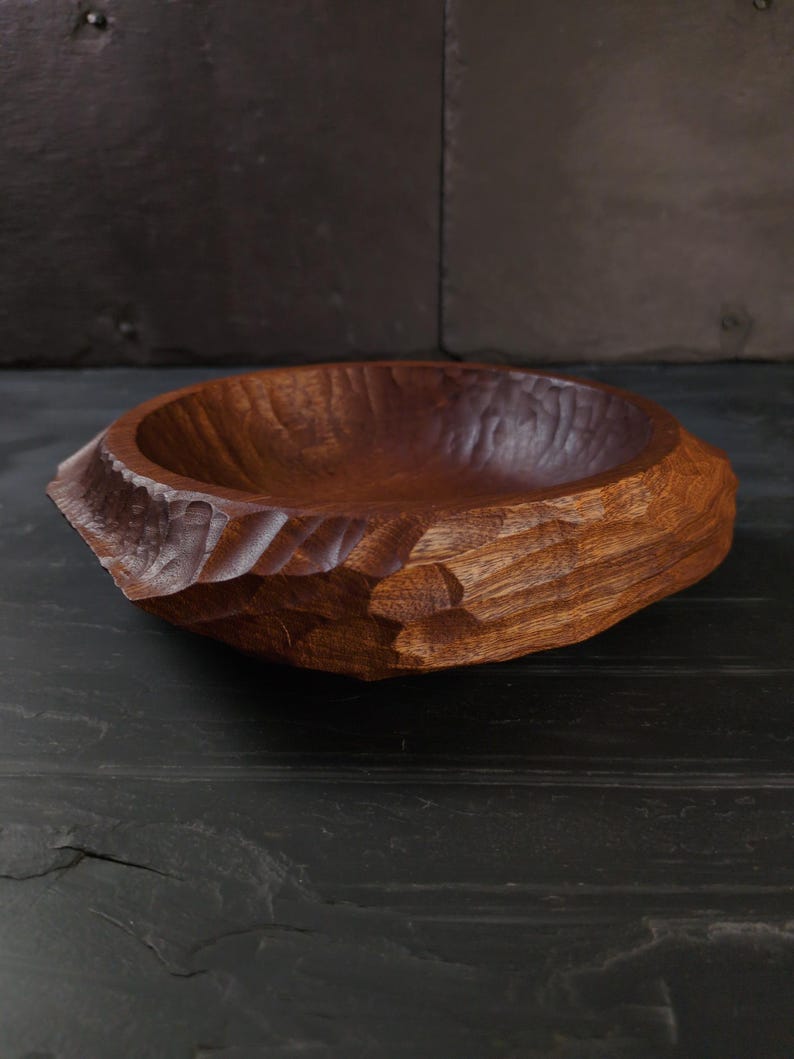 May include: A hand-carved wooden bowl. The bowl has a rough, textured exterior and a smooth interior. The bowl is a rich, warm brown colour, showcasing the natural wood grain. The bowl is likely used for serving or display.