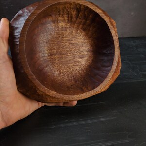 May include: A hand-carved wooden bowl with a natural, dark brown finish. The bowl has a rough, textured exterior and a smooth, rounded interior. The wood grain is visible, adding to the rustic aesthetic. The bowl is held by a hand.