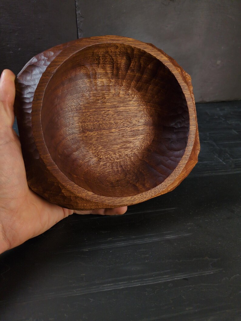 May include: A hand-carved wooden bowl with a natural, dark brown finish. The bowl has a rough, textured exterior and a smooth, rounded interior. The wood grain is visible, adding to the rustic aesthetic. The bowl is held by a hand.