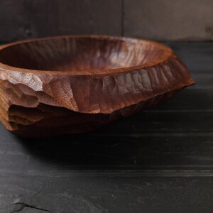 May include: A dark brown, hand-carved wooden bowl. The exterior has a textured, faceted design, while the interior is smooth. The wood grain is visible. This bowl is likely for serving or display. The bowl's dimensions are in centimetres.