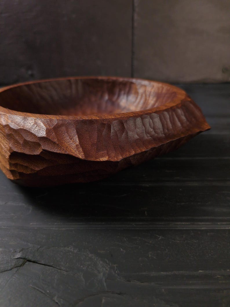May include: A dark brown, hand-carved wooden bowl. The exterior has a textured, faceted design, while the interior is smooth. The wood grain is visible. This bowl is likely for serving or display. The bowl's dimensions are in centimetres.