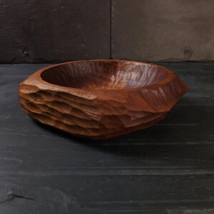 May include: A hand-carved wooden bowl with a textured exterior and a smooth interior. The bowl is a rich brown colour and appears to be made from a single piece of wood. The bowl is on a dark surface.